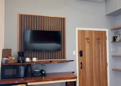 Kitchenette bar with stools and shelf showing microwave, mini fridge, wine glasses and coffee maker, as well as complimentary tea and coffee. Table can be pulled out from bar for extra space and above the bar is a wall munted TV.